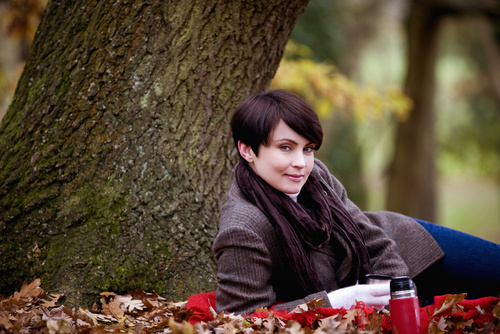 Beautiful woman with short, brown hair in pants and brown jacket lying in front of tree with leaves, fall setting